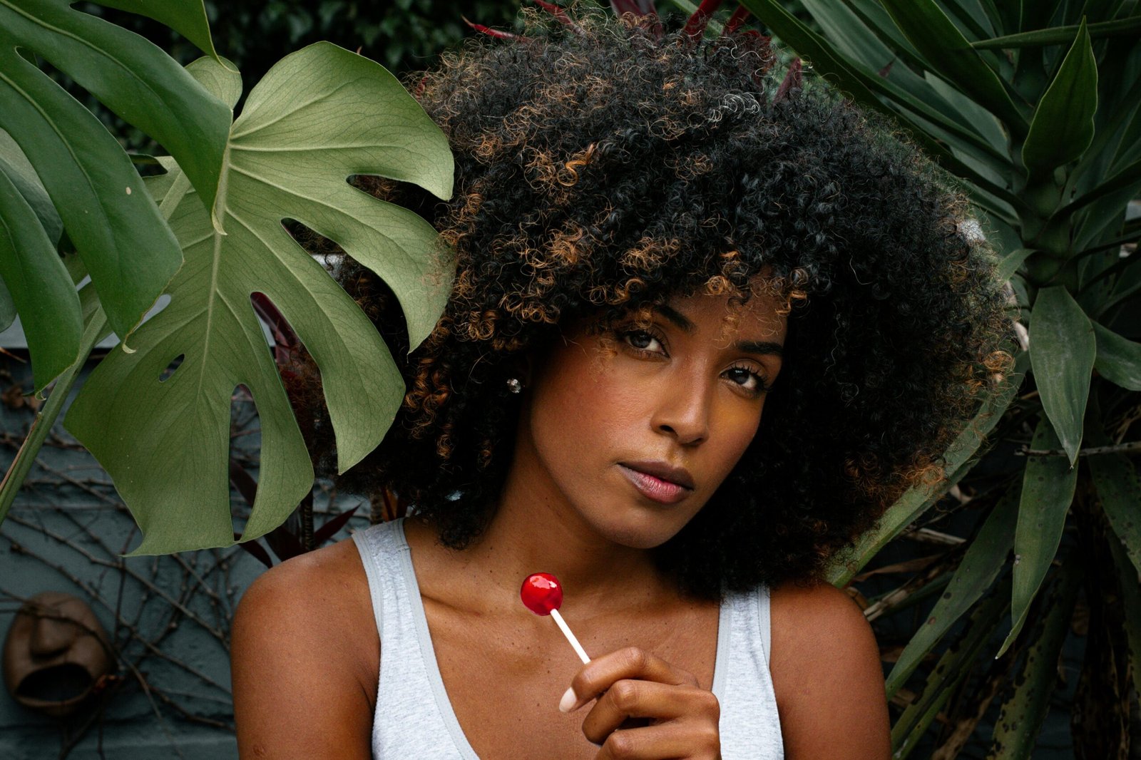 A woman with afro-textured hair holds a lollipop outdoors, surrounded by tropical plants.